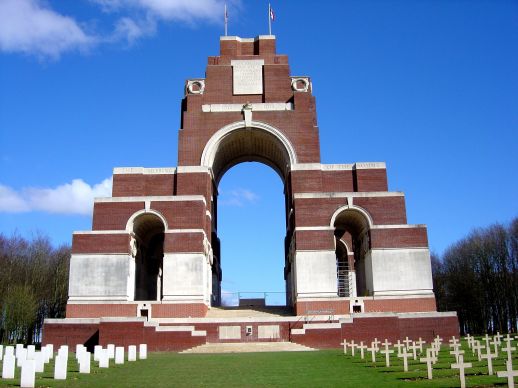 Photo of Thiepval Memorial. Red brick and white stone memorial with 3 arches. Rows of headstone to left front. Rows of cross marker to the right front.