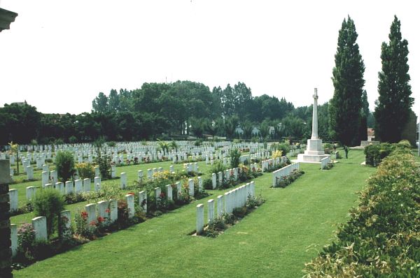 Photo of Menin Road South Cemetery. Rows of headstones and plants with a white stone cross memorial to the right with trees in the background.