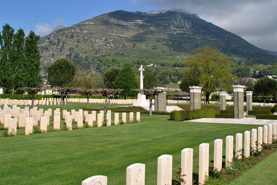 Rows of white gravestones with plants in front of them between areas of mown grass. The mountain of Monte Cassino is prominent in the background