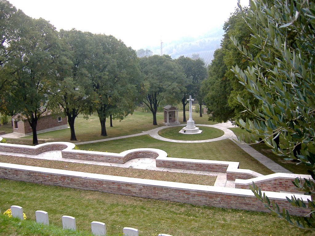 a brick structure on the side of the hill with, at the bottom, surrounded by trees is the white cross of sacrifice