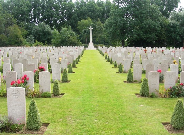 Rows of white gravestones with plants in front of them. A mown strip of grass leads up to the Cross of Sacrifice in the distance with trees beyond