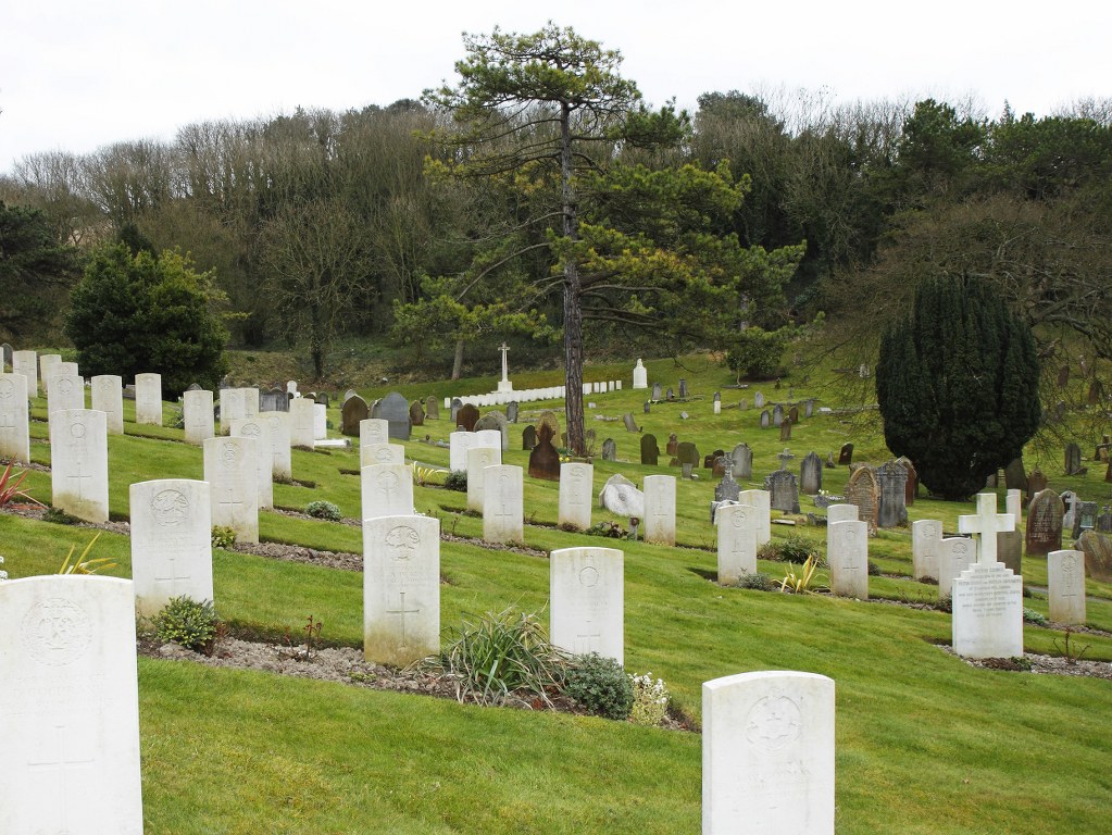 rows of white crosses  on the hillside between mown grass , the bottom of the valley is visible with the shite cross of sacrifice at the bottom and trees on the other side of the valley