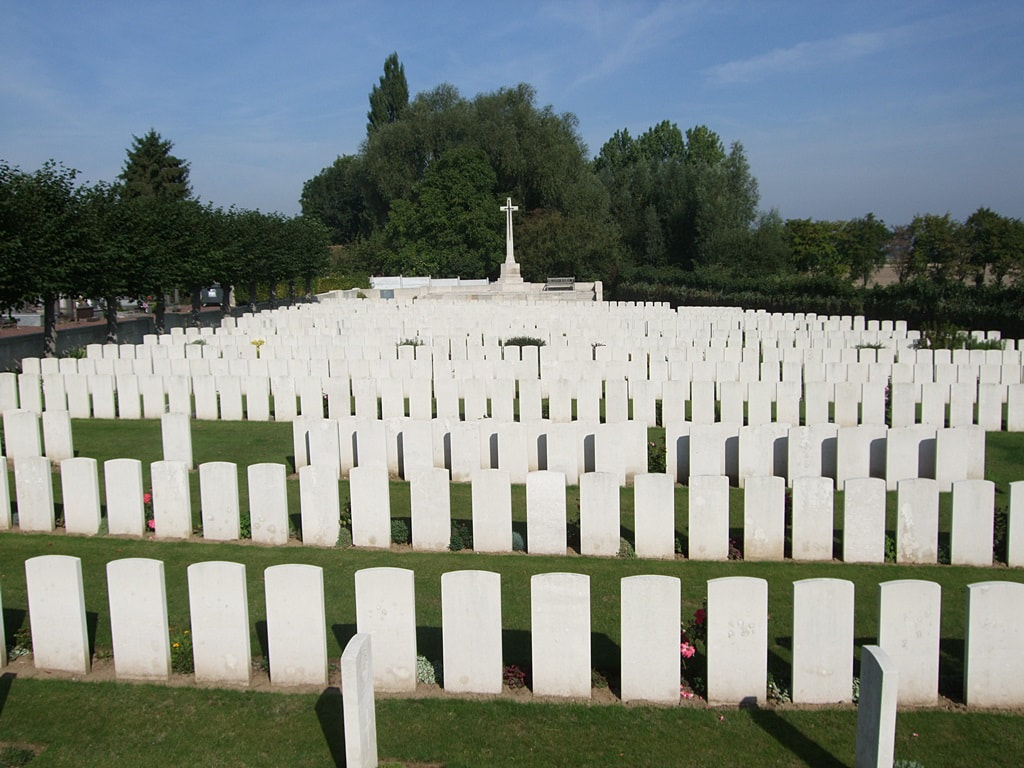 Rows of white gravestones with the white Cross of Sacrifice in the distance. The cemetery is surrounded by trees
