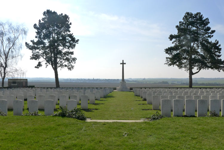Photo of Highland Cemetery Le Cateau. Rows of white headstones either side of a grass path leading to a cross monument with a tree on both sides.