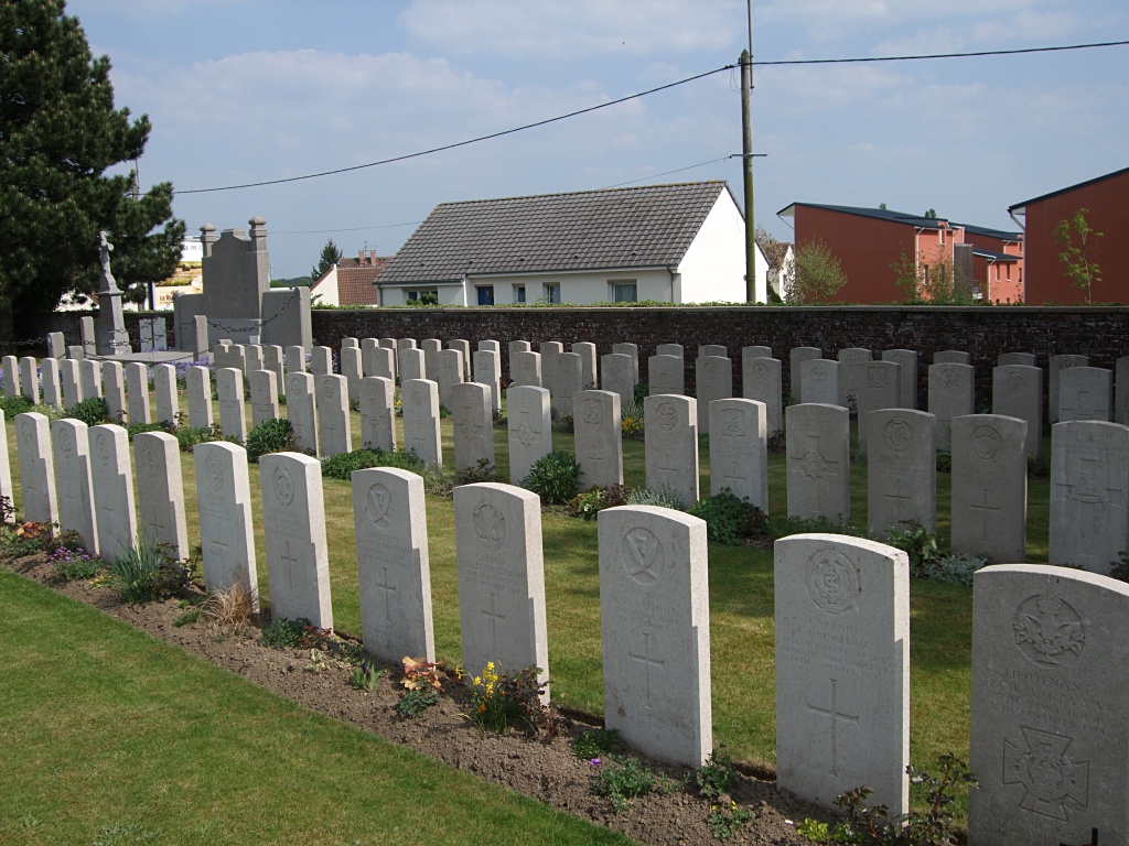 Rows of white gravestones with plants in front of them between strips of mown grass. Houses can be seen beyond the graveyard