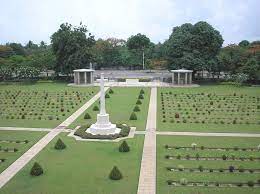 Photo of Taukkyan War Cemetery. Grass lawn with paths and a white stone cross monument in the middle.