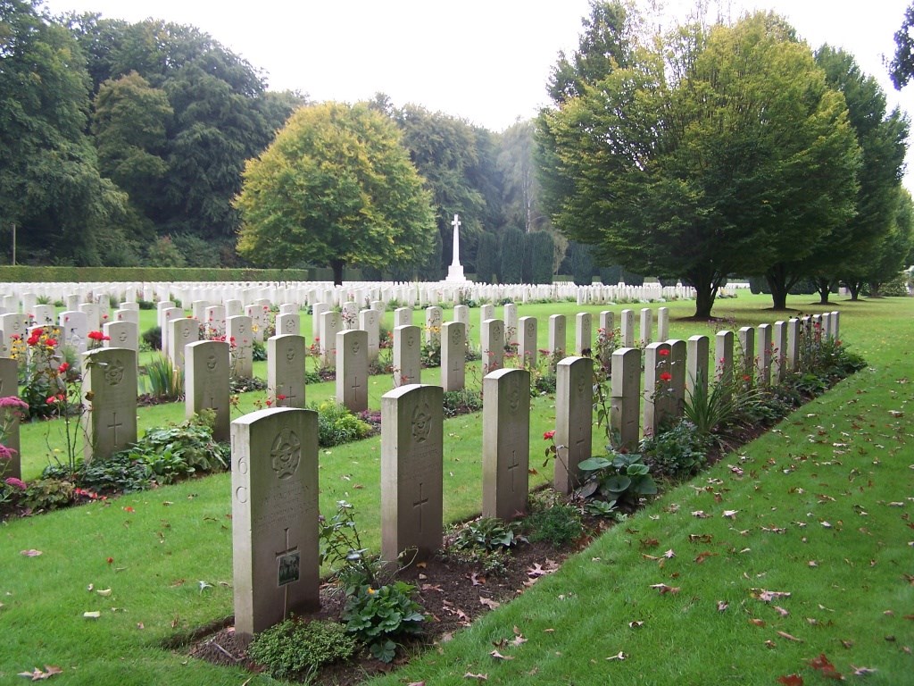 rows of headstones in mown grass with the white cross of sacrifice in the background