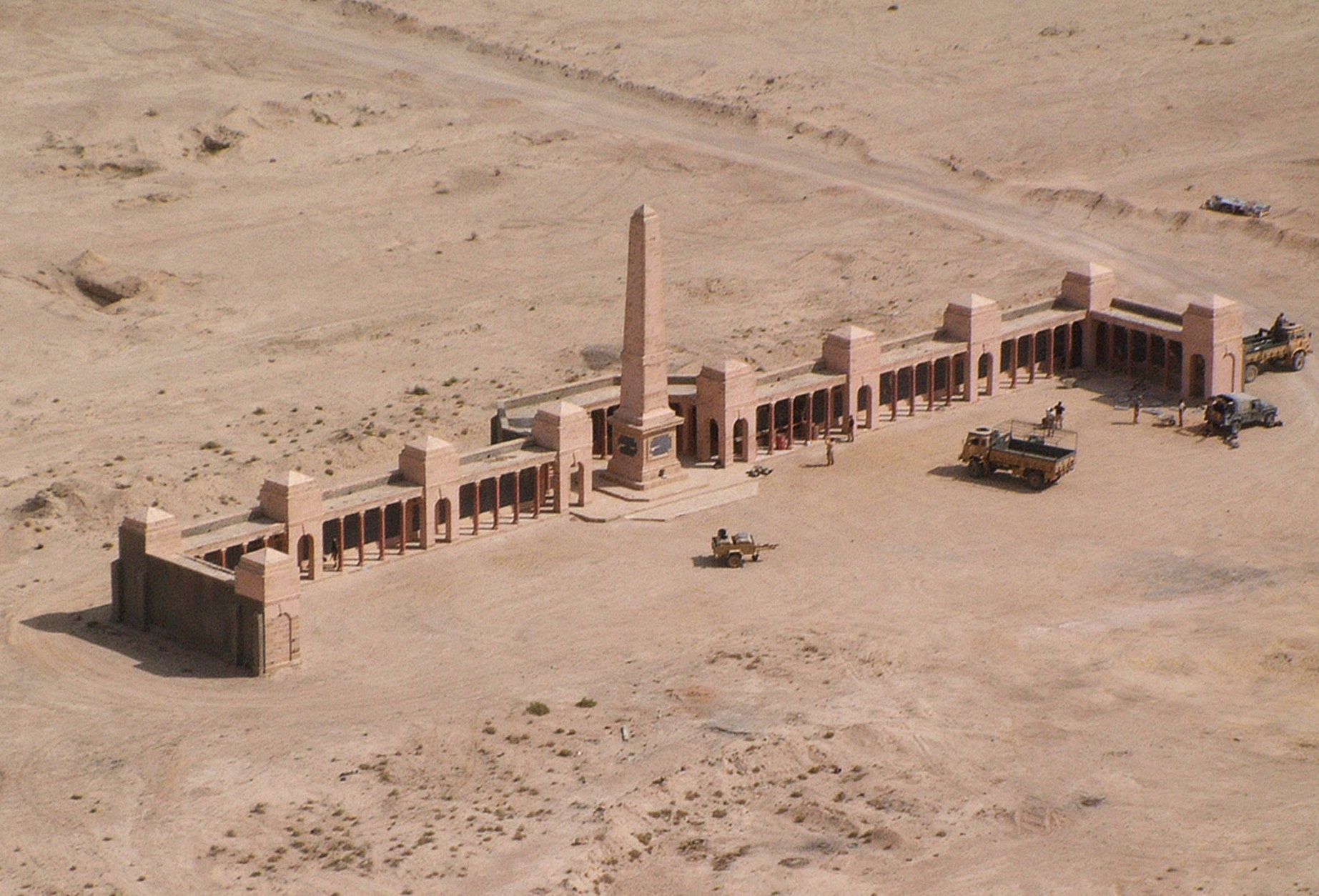 Photo of Basra Military Cemetery. Sand ground with a long wall memorial with 11 towers. The central tower is the tallest. 3 military vehicles and a trailer are in front,