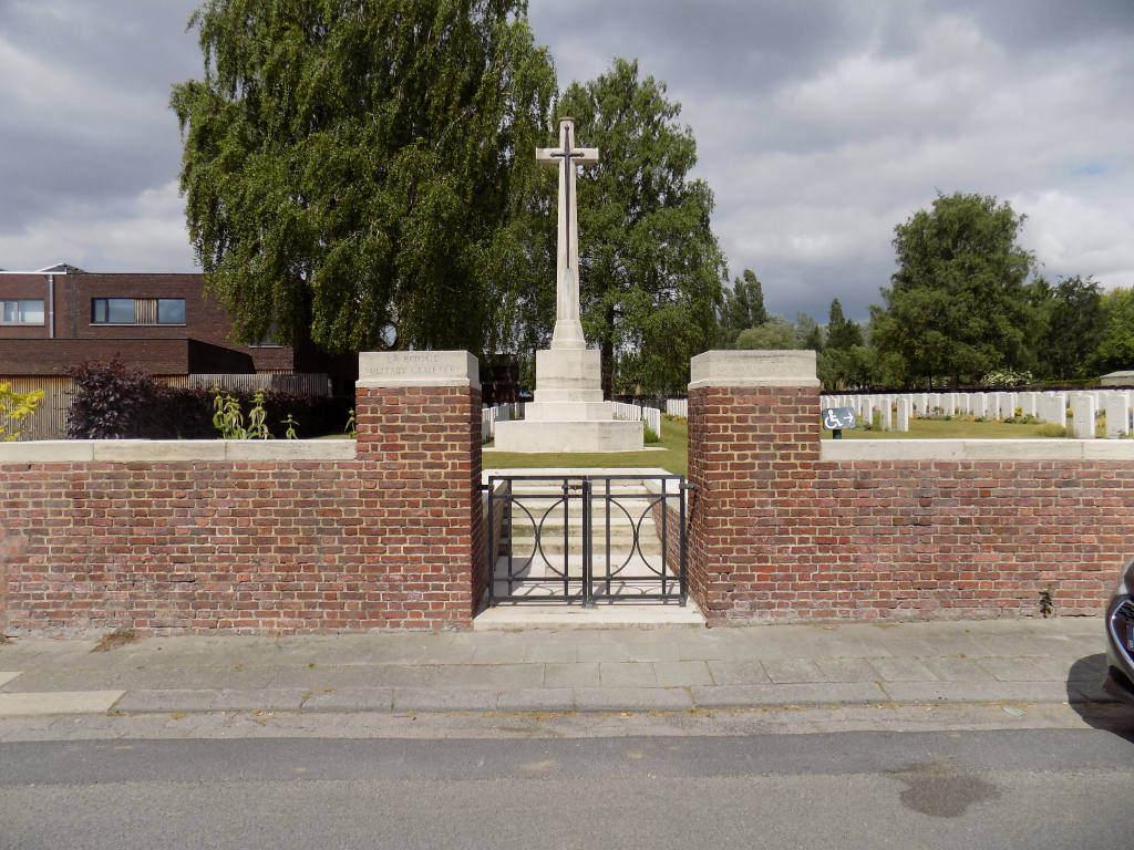 Photo of La Brique Military Cemetery. Red brick wall in front of rows of headstones a cross memorial.