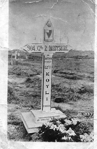 the original white cross marking his grave which gives his details. some flowers are placed at the foot of the grave