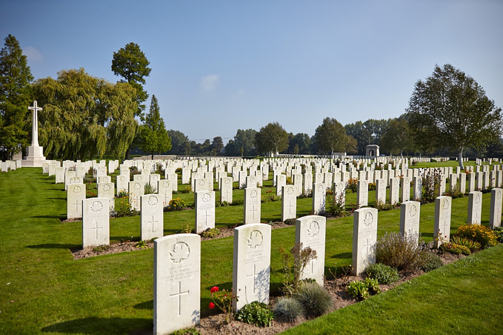 Photo of Railway Dugouts Burial Ground. Rows of headstones and plants in front of a cross memorial and trees.