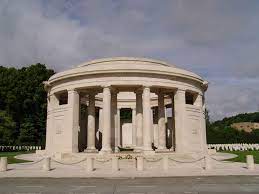 Photo of Ploegsteert Memorial. A circular memorial of columns in front of headstones and trees.