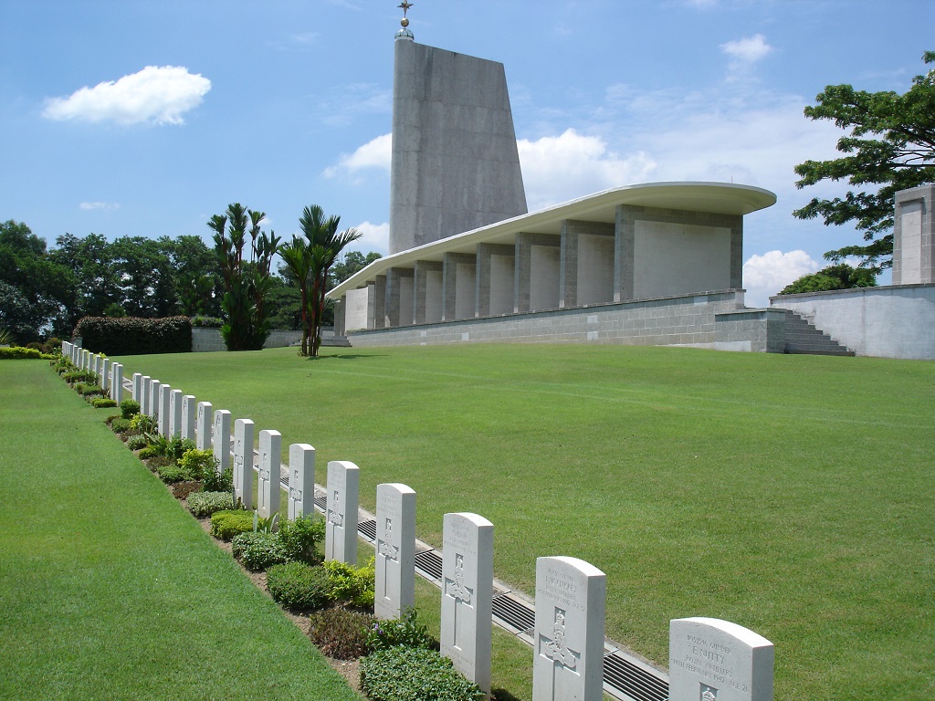 a row of white crosses at the bottom of the hill with a modern white building on the hill above