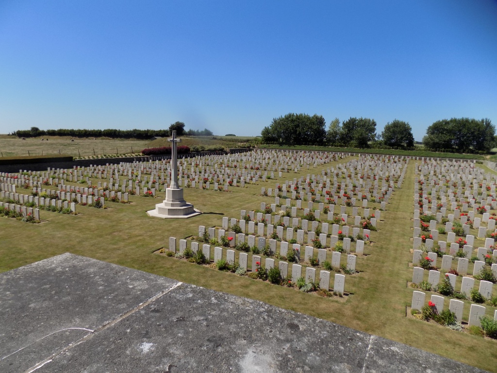 Looking down into a graveyard with rows of white gravestones and plants in front of them. Areas of mown grass are between the rows and the white Cross of Sacrifice can be seen