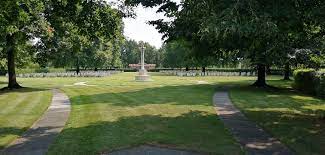 a white cross stands in a grass field with rows of white gravestones in the background with trees