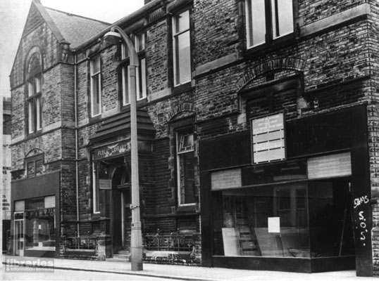 A black and white photograph of the public library block, Station Road, Ossett, shortly before its cleaning and modernisation.  Empty shops sit either side of the building.  Internal Reference: OS 23