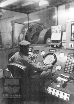 A black and white photograph of part of the Control Room at Glasshoughton Colliery, Glasshoughton, Castleford.  A man is at the control panel. The colliery closed in 1986.  Through the glass screen machinery can be seen.  Internal Reference: CA YILLUS 4510