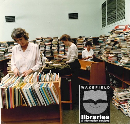 A colour photograph of staff working in the Circulation Department at Wakefield Library Headquarters, Balne Lane, Wakefield. Bernie Metcalfe is on the left, Ann Haith on the right. The piles of books on the counter are known as "the hump" and are waiting to be recirculated to other libraries. Taken September 1991. Internal Ref: WF Local Studies Library
