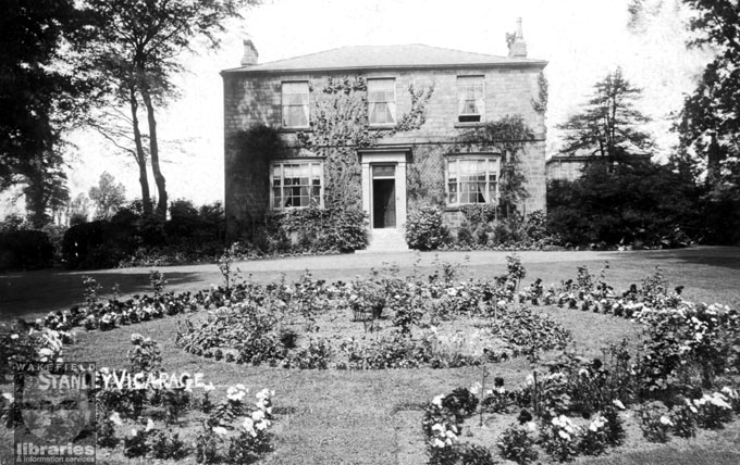 A black and white postcard showing the vicarage of Saint Peter's Church, Aberford Road, Stanley.  The vicarage is a double fronted house with a lawn and flower beds at the front.  Internal Reference: WR (LS)