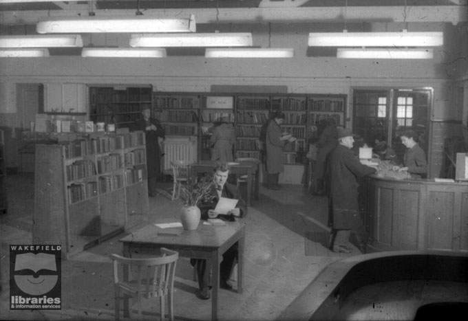 Wakefield Drury Lane Library after alterations, Wakefield, 1956 A black and white slide showing the interior of Drury Lane Library, Wakefield, in 1956, after alterations to the layout to make the library more open and welcoming. The bookshelves were lowered, the counter has been moved to a more accessible position near the entrance doors and tables and chairs have been provided for borrowers' use. Internal Reference: WR (LS)
