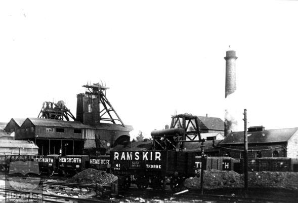 A black and white photograph showing Hemsworth Colliery.  The pit headgear can be seen to the left and the boilerhouse chimney is to the right.  The railcars that hold the coal have 'Hemsworth' or 'Ramskir' painted on them.  The pit was sunk between 1876 and 1877 and was finally closed in 1967. The land has since been redeveloped.